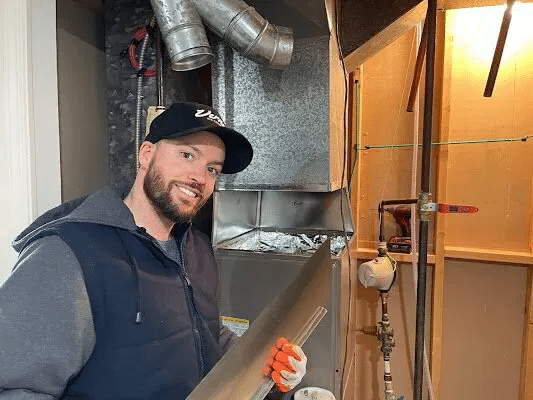 A man smiles while holding a pipe in a bathroom, representing Vernon AC staff's friendly service.