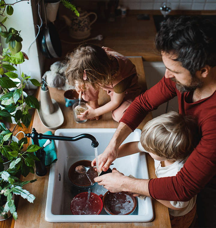 Dad, daughter, and son washing dishes together at the kitchen sink, sharing smiles and teamwork in a family setting.
