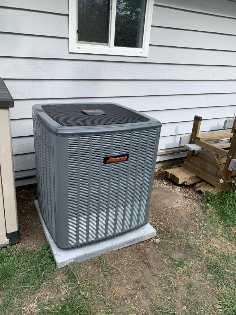 Heat pump outside a house, featuring an Amana System, designed for efficient heating and cooling.