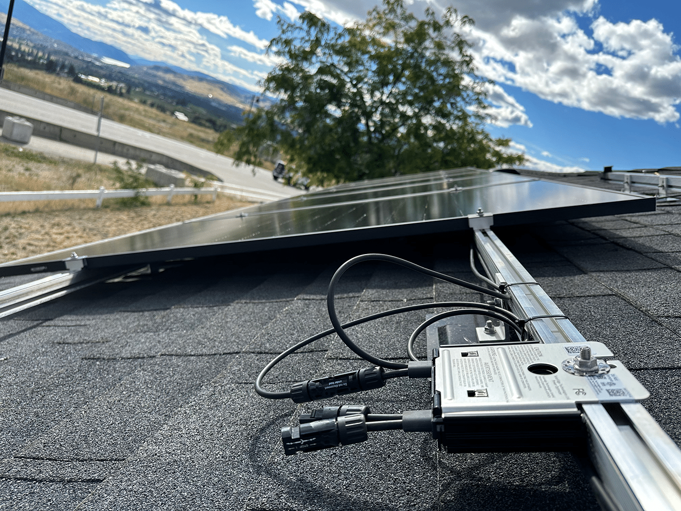 A close-up view of a solar panel mounted on a roof, with electrical connections and a background of hills and clouds.