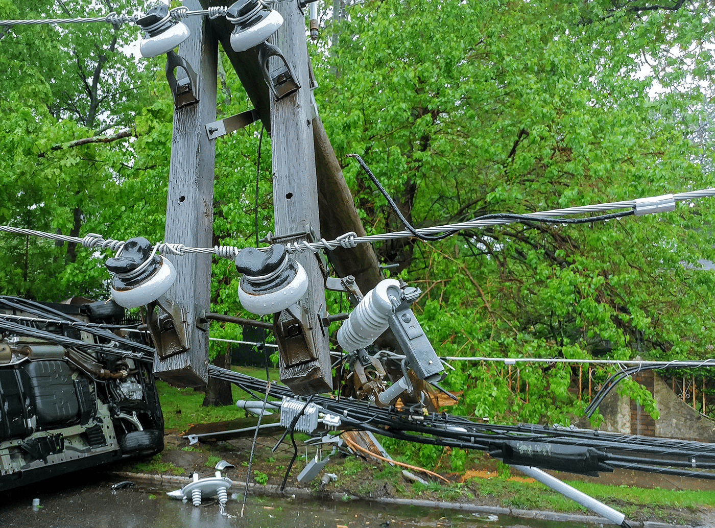 A damaged utility pole with power lines and insulators, surrounded by greenery, and an overturned vehicle in the background.