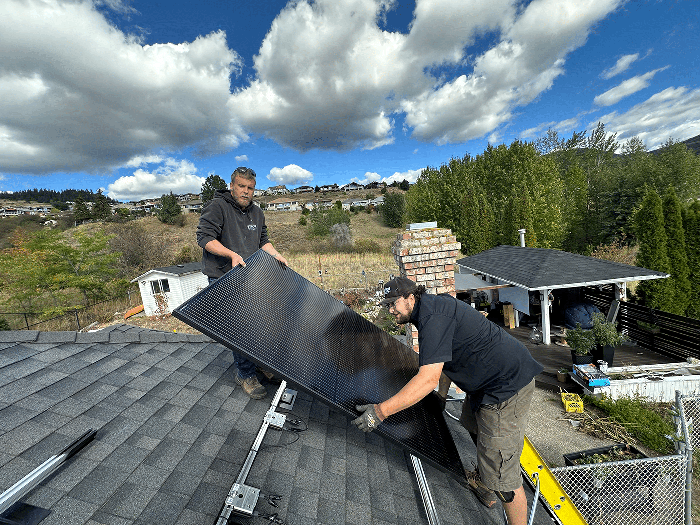 Two workers are installing a solar panel on a roof, with a residential area and trees visible in the background under a partly cloudy sky.