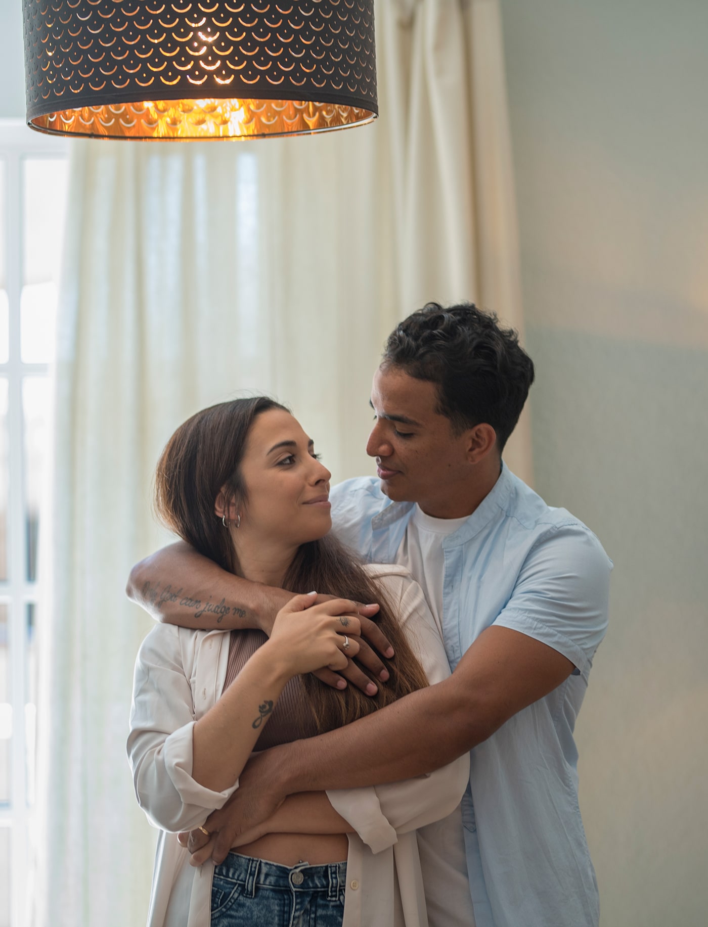 Young millennial interracial couple hug with love at home in living room - relationship with black boy and caucasian girl together standing and embracing looking each other - concept of life and house