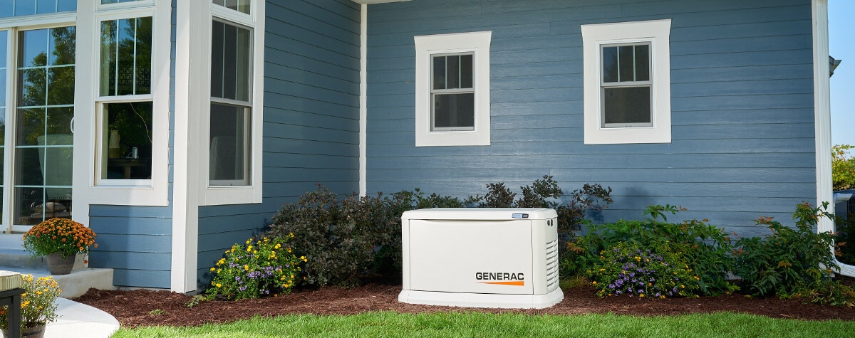 A white Generac generator next to a blue house with two windows and landscaped flowers.