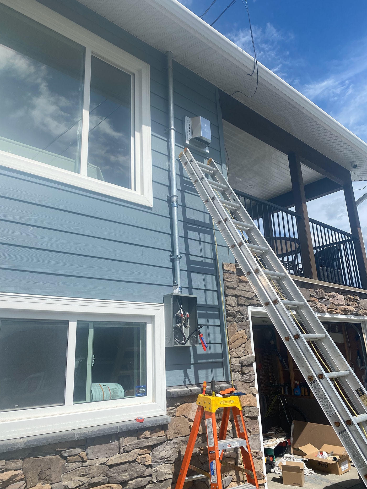A blue house exterior with a ladder leaned against it, a mounted electrical box, and a small orange step ladder nearby.
