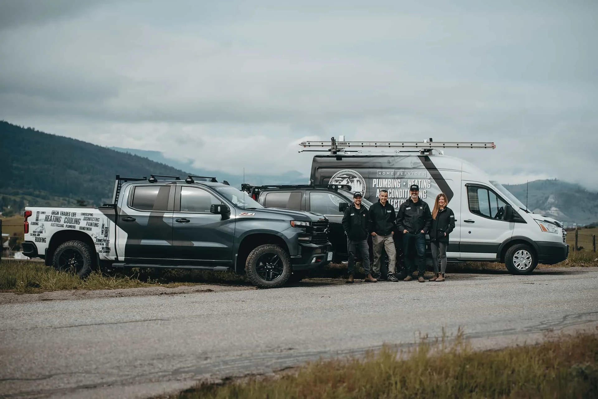 Three men in work attire and a woman stand beside their trucks, with the Okanagan majestic mountains in the background, representing Vernon Air Conditioning, Plumbing & Electrical Services Team Staff.